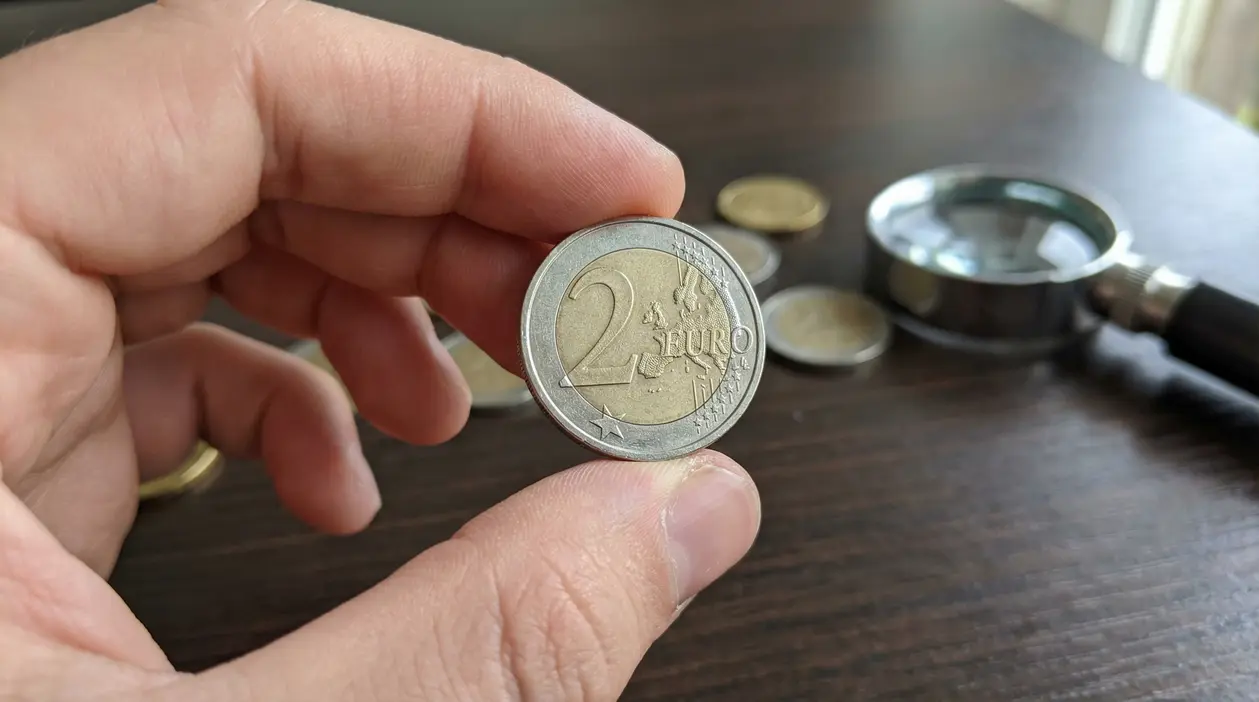 Mano che tiene una moneta da 2 euro con altre monete e lente d'ingrandimento sullo sfondo