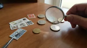 Una mano tiene una lente di ingrandimento sopra monete e francobolli su un tavolo in legno.