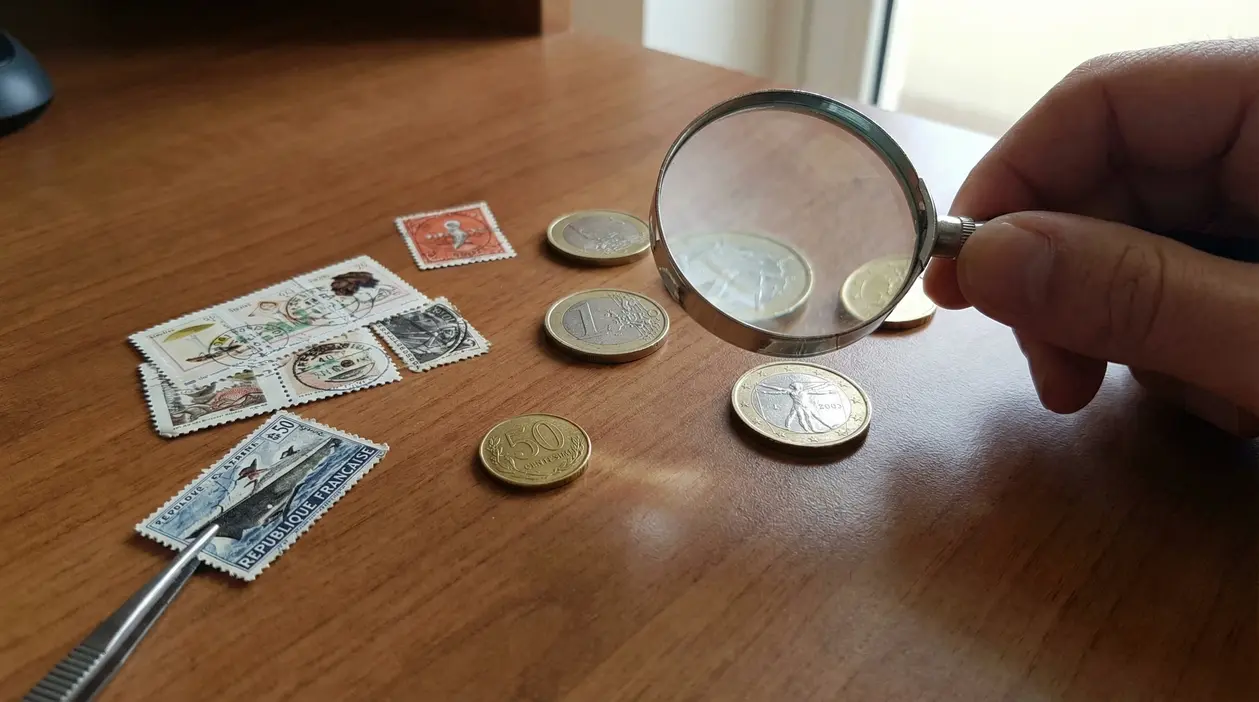 Una mano tiene una lente di ingrandimento sopra monete e francobolli su un tavolo in legno.
