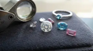 Varie gemme colorate e un anello esposti su un panno grigio vicino a una lente da gioielliere