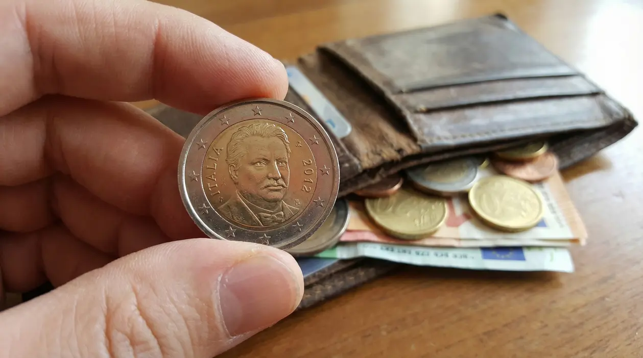 Moneta da 2 euro con volto inciso tenuta in mano davanti a un portafoglio aperto con altre monete e banconote.