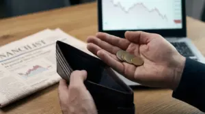 Una persona tiene alcune monete accanto a un portafoglio vuoto, con un grafico di borsa in calo sullo sfondo.