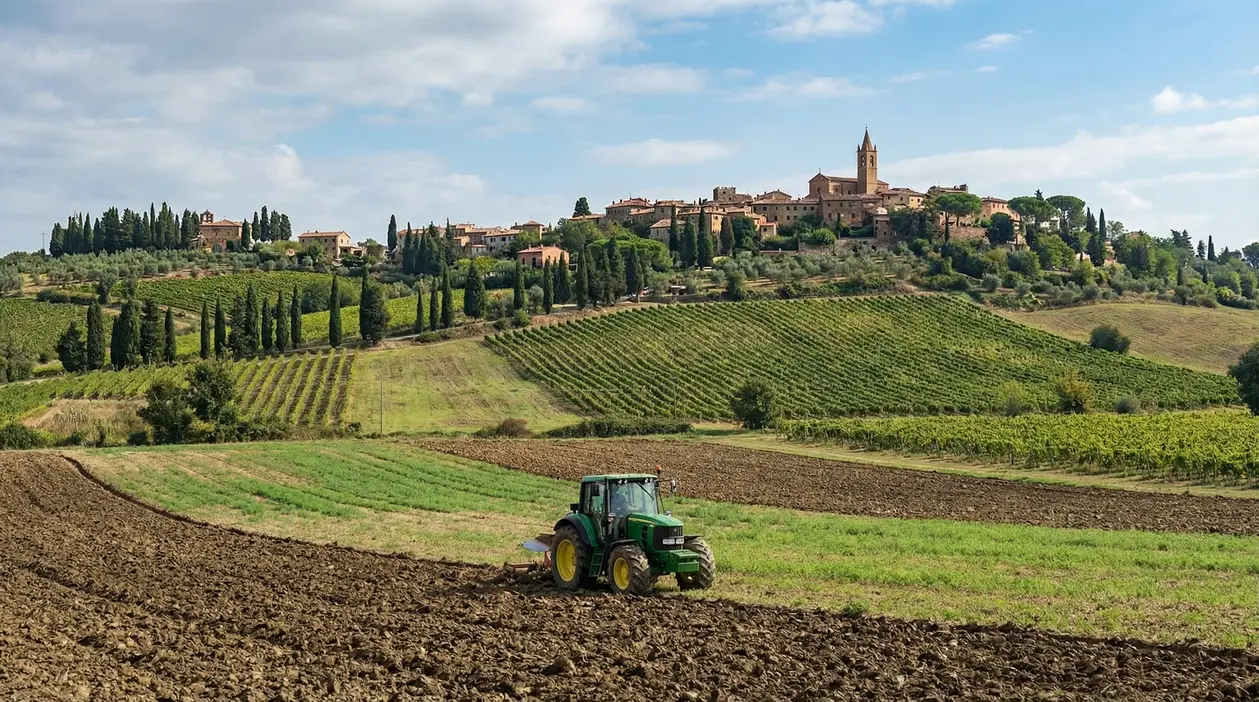 Trattore al lavoro in un terreno agricolo con un borgo collinare sullo sfondo