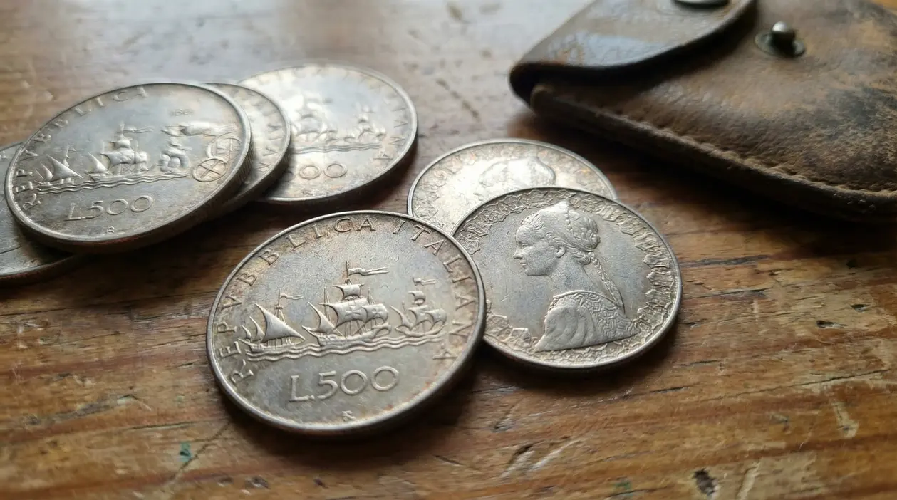 Vecchie monete italiane da 500 lire d’argento appoggiate su un tavolo di legno accanto a un portamonete in pelle.