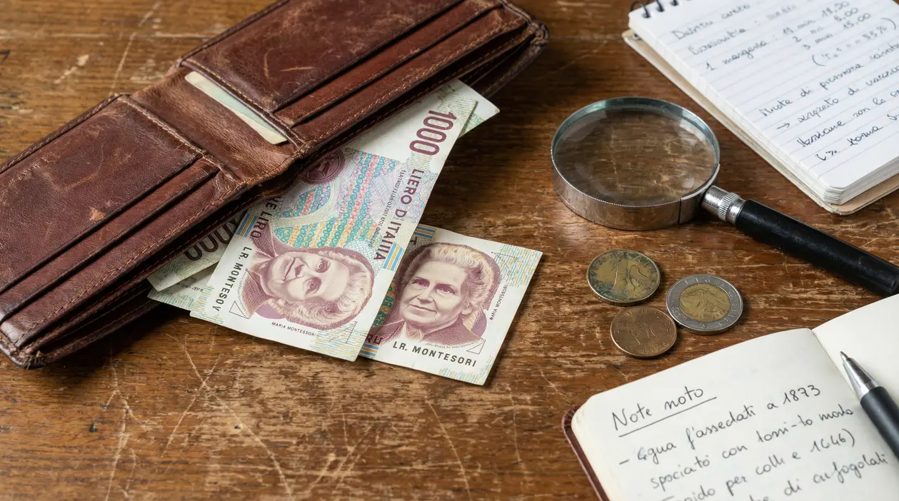 Banconote da mille lire con Maria Montessori accanto a un portafoglio di pelle e alcune monete su un tavolo