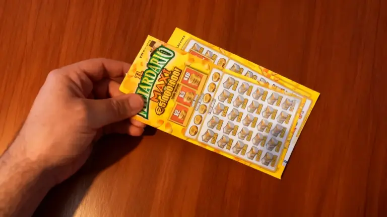 Una mano tiene in primo piano due biglietti del gratta e vinci su un tavolo di legno